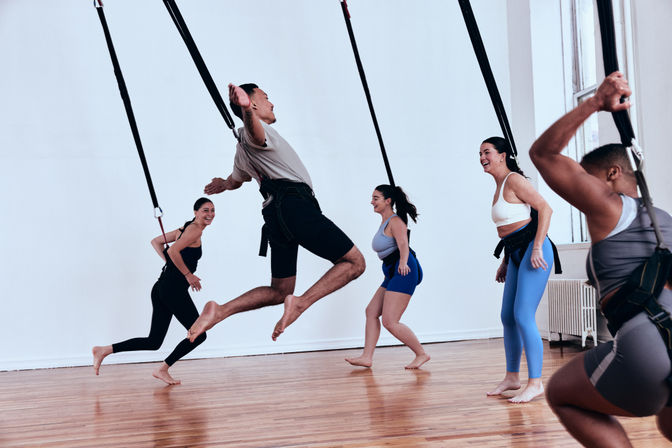 Adults in a bright indoor fitness studio doing an aerial suspension class — one person mid-jump while others laugh and practice moves on a hardwood floor.