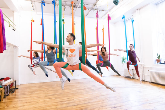 Group aerial yoga class in a bright fitness studio, adults suspended in colorful silk hammocks doing wide‑leg poses with arms extended over a hardwood floor.