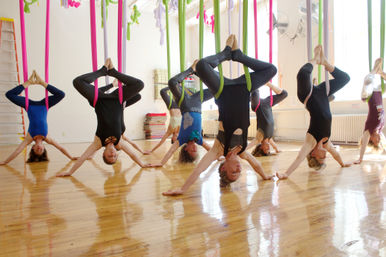 Group aerial yoga class with students suspended upside-down in colorful silk hammocks (pink and green), performing inversion poses over a bright hardwood-floored studio.