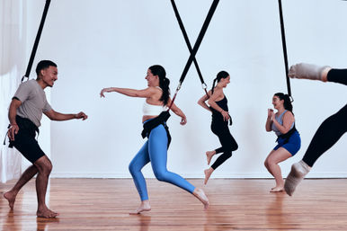 Energetic group bungee fitness class in a bright indoor studio, adults wearing harnesses attached to ceiling straps, laughing and jumping over a wooden floor.