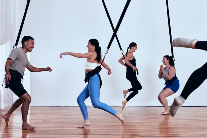 Energetic group bungee fitness class in a bright indoor studio, adults wearing harnesses attached to ceiling straps, laughing and jumping over a wooden floor.