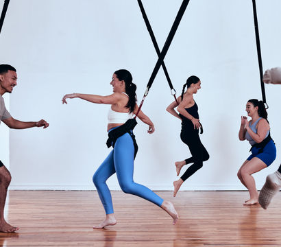 Energetic group bungee fitness class in a bright indoor studio, adults wearing harnesses attached to ceiling straps, laughing and jumping over a wooden floor.