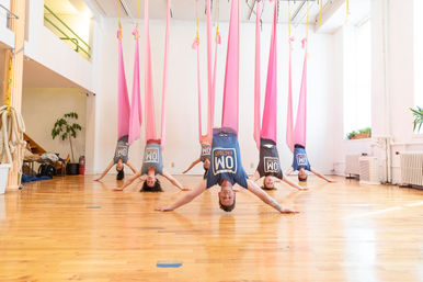 Bright loft yoga studio with five participants suspended upside-down in pink aerial hammocks, arms outstretched on a polished hardwood floor during a playful aerial yoga class