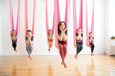 Five people in a bright aerial yoga class suspended in pink silk hammocks over a hardwood studio floor, smiling and posing with hands under their chins.