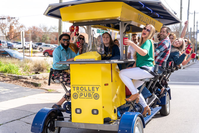 Group of friends laughing and toasting drinks on a bright yellow pedal pub trolley cruising an urban street on a sunny day.