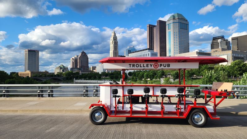 Red-and-white pedal pub trolley with canopy and bar seats parked on a bridge over a river, framed by a sunlit downtown skyline and puffy blue clouds — urban riverfront scene.