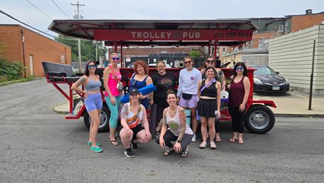 Group of smiling women in colorful athletic wear and sunglasses posing with yoga mats and water bottles in front of a red open-air pedal trolley parked on an urban side street.