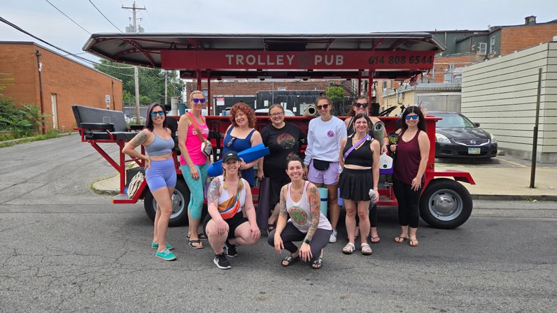 Group of smiling women in colorful athletic wear and sunglasses posing with yoga mats and water bottles in front of a red open-air pedal trolley parked on an urban side street.