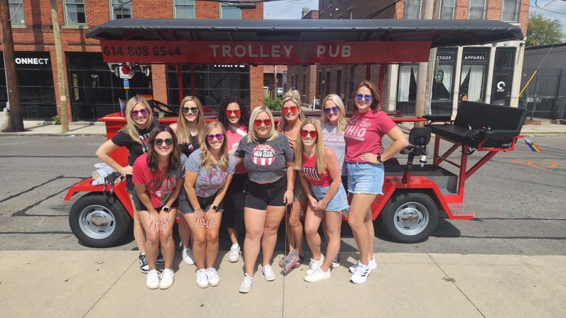 Group of women in sunglasses and casual Ohio-themed tees posing in front of a red open-air party trolley on a sunny downtown street with brick storefronts.