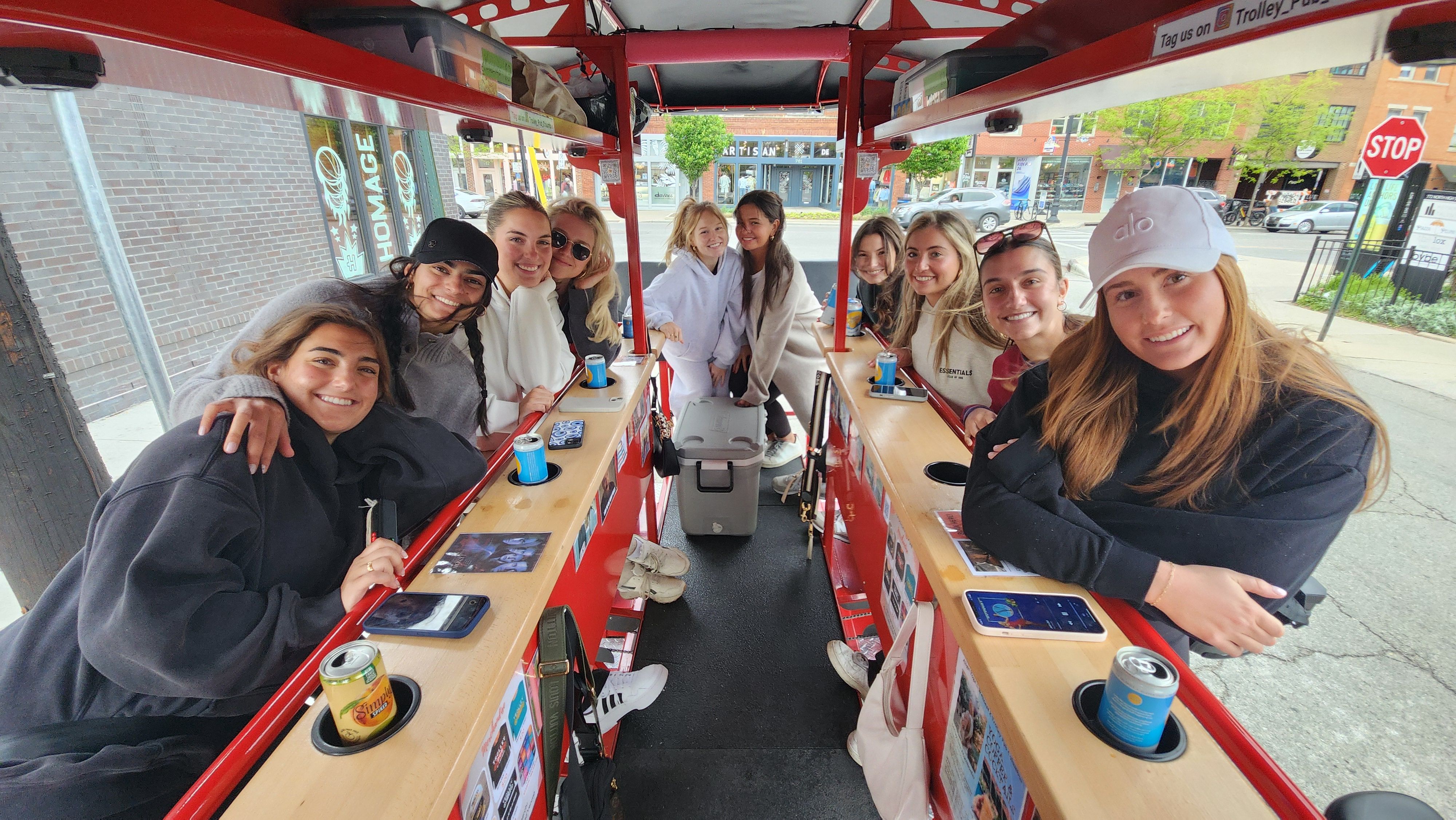 Smiling group of friends riding a red pedal-powered party bike on a small downtown street, seated along wooden bars with drinks and phones for a daytime city tour vibe.