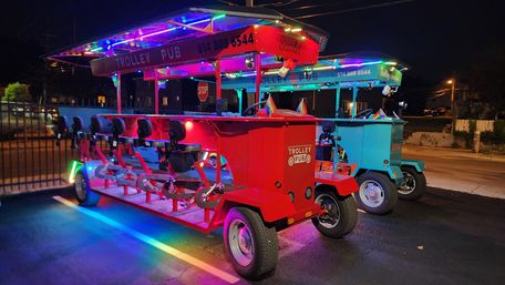 Neon-lit red and teal pedal-powered party trolleys parked on an urban street at night, open-air bar-style seats and pedals under a festively lit canopy