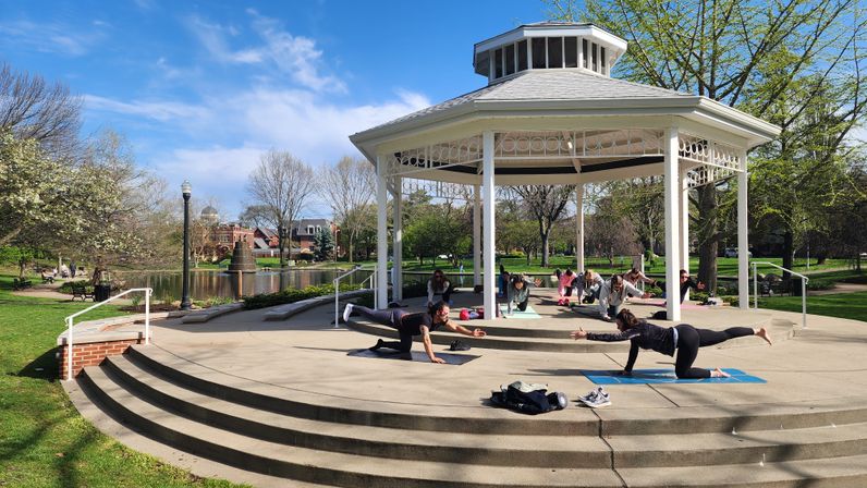 Community outdoor yoga class at a white park gazebo beside a pond, participants on mats stretching on a sunny spring day with blossoming trees and walking paths