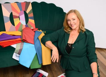 Smiling woman in a green blazer seated beside a green velvet couch draped with bright fabric swatches and a colorful color-wheel board — color styling setup.