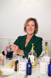 Smiling woman in a green blazer blending blue liquid at a marble counter surrounded by glass pump bottles, dropper bottles and scent testing strips in a boutique perfume and skincare studio