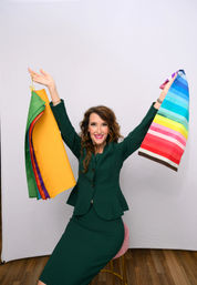 Smiling woman in an emerald green suit seated on a pink stool in a photo studio, holding up bright fabric swatches and a rainbow-striped textile