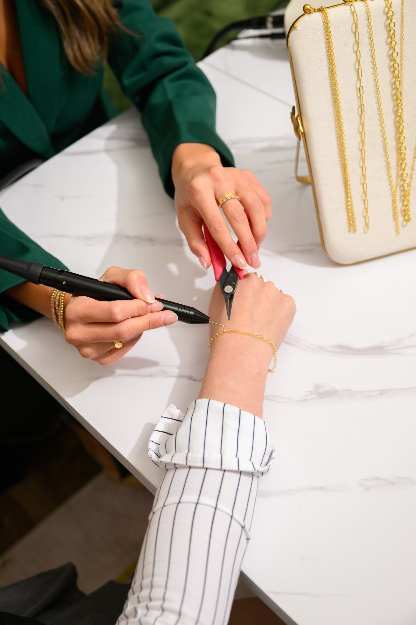 Jeweler repairing a delicate gold chain bracelet on a customer's wrist with pliers and a precision tool at a white marble counter, gold necklaces displayed nearby.