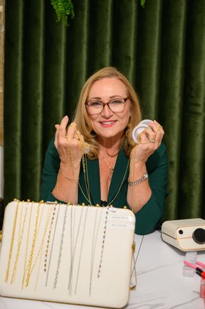 Smiling woman with glasses holding gold chain necklaces and spools over a tabletop jewelry display of necklaces, set against a green velvet curtain backdrop