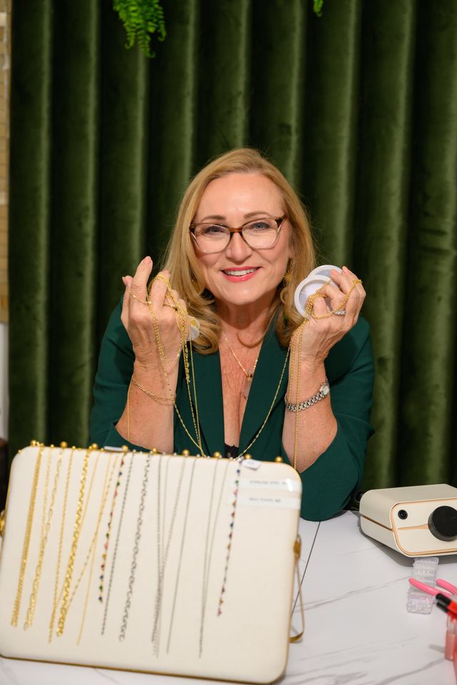 Smiling woman with glasses holding gold chain necklaces and spools over a tabletop jewelry display of necklaces, set against a green velvet curtain backdrop