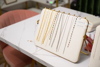 White padded display rack holding gold and silver chain necklaces with small multicolored gemstone accents on a marble table, pink velvet chair in the background.