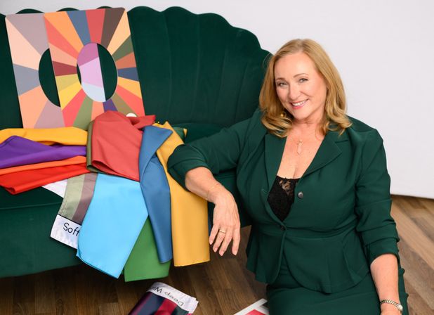 Smiling woman in a green suit beside a dark green velvet sofa draped with vivid fabric swatches and a multicolor seasonal palette—personal color consultation and styling setup.