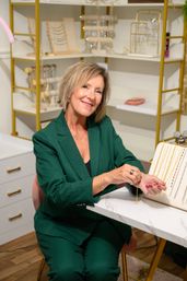 Smiling woman in a green blazer trying on a gold bracelet at a boutique jewelry display with necklaces and organized shelving in the background.