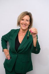 Smiling professional woman in a green blazer holding a gold chain necklace with a black pendant against a plain white backdrop.