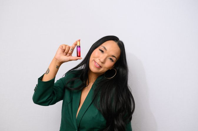 Smiling person with long black hair wearing a green blazer and hoop earring holding a small pink-to-purple cosmetic bottle with a gold cap against a plain white background