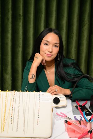 Jewelry designer at a worktable — person in a green blazer and hoop earrings poses behind a display of delicate necklaces and beaded chains with pliers, tools, and an organizer on a white table against a green velvet curtain backdrop.