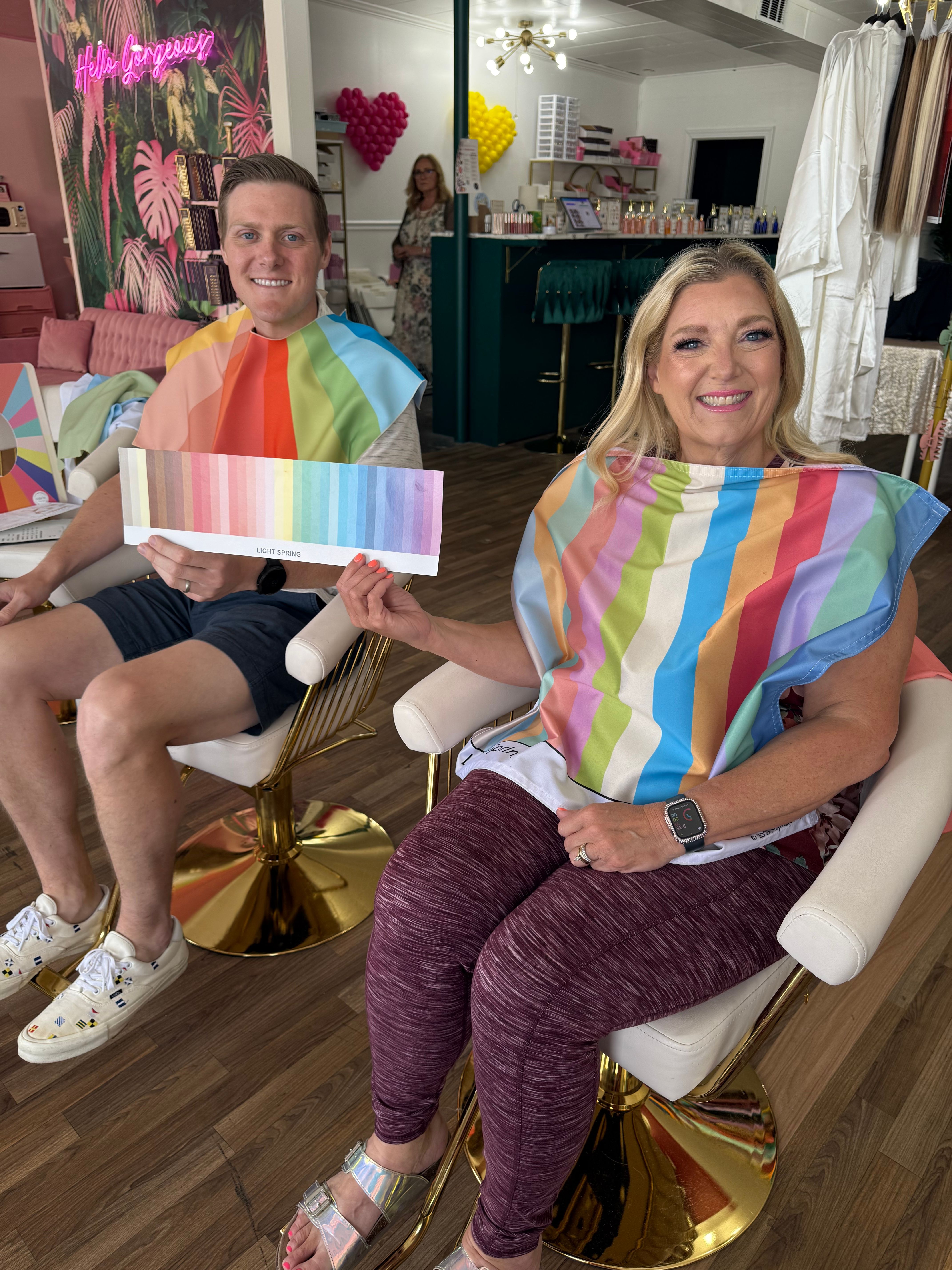 Smiling man and woman in a boutique salon during a color analysis session, wearing bright seasonal drapes and holding a “Light Spring” palette.