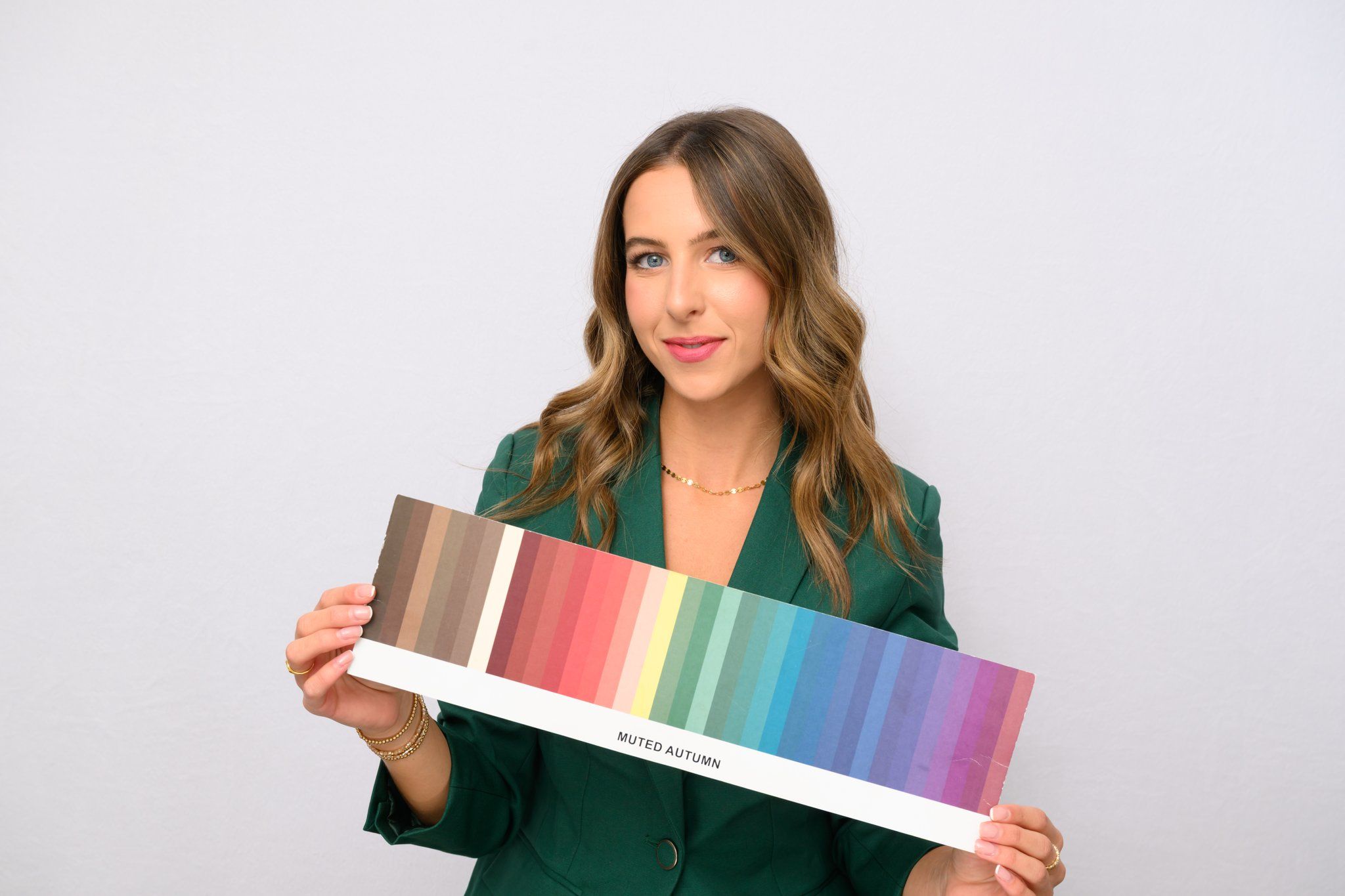 Studio portrait of a smiling woman in a green blazer holding a 'Muted Autumn' color swatch palette showing warm-to-cool seasonal hues — color analysis and styling reference