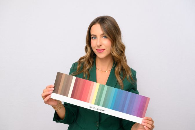 Studio portrait of a smiling woman in a green blazer holding a 'Muted Autumn' color swatch palette showing warm-to-cool seasonal hues — color analysis and styling reference
