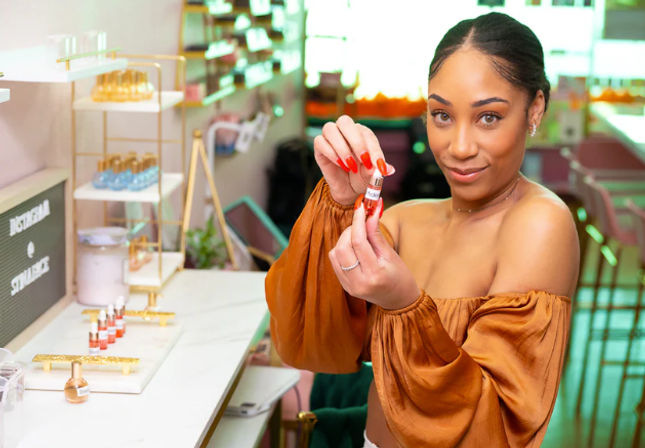 Smiling woman in an off-shoulder rust top holding a red lipstick at a bright cosmetics counter in a trendy beauty boutique with makeup displays on shelves
