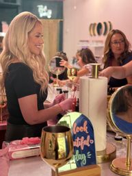 Smiling blonde woman in a pink-themed beauty salon getting a manicure from a technician in pink gloves, surrounded by mirrors, gold wine glasses and a decorative "look beautiful" sign.