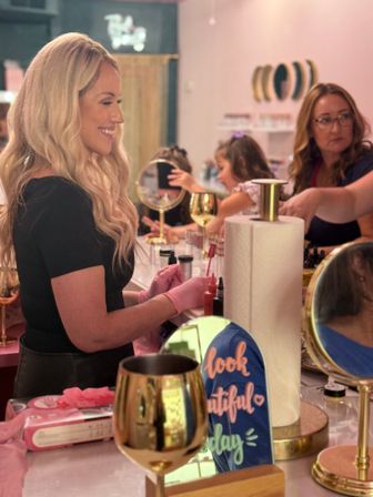 Smiling blonde woman in a pink-themed beauty salon getting a manicure from a technician in pink gloves, surrounded by mirrors, gold wine glasses and a decorative "look beautiful" sign.