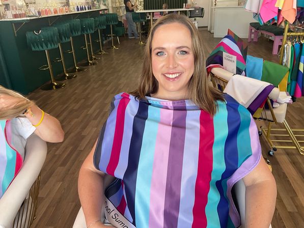 Smiling woman seated in a color-consultation salon wearing a bright multicolored striped drape, surrounded by fabric swatches, green bar stools and wood-look flooring.