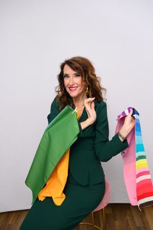 Studio portrait of a smiling woman in a green dress seated on a pink stool holding colorful fabric swatches and scarves for a personal color consultation