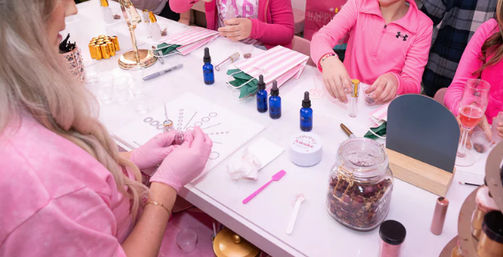 Pink-themed DIY fragrance workshop: an adult in pink gloves guides children around a craft table with blue dropper bottles, test tubes, a jar of dried flowers, spatulas and striped napkins at a birthday-style party.