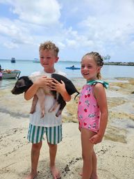 Two children on a tropical sandy beach holding a black-and-white piglet, with clear turquoise water, anchored boats and a partly cloudy sky in the background.