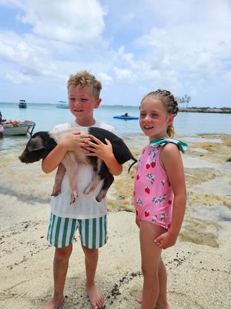 Two children on a tropical sandy beach holding a black-and-white piglet, with clear turquoise water, anchored boats and a partly cloudy sky in the background.