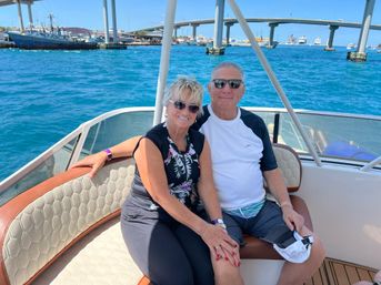 Smiling couple in sunglasses seated on a boat bench on a sunny day, with turquoise harbor water, a coastal bridge and cruise ships in the background.