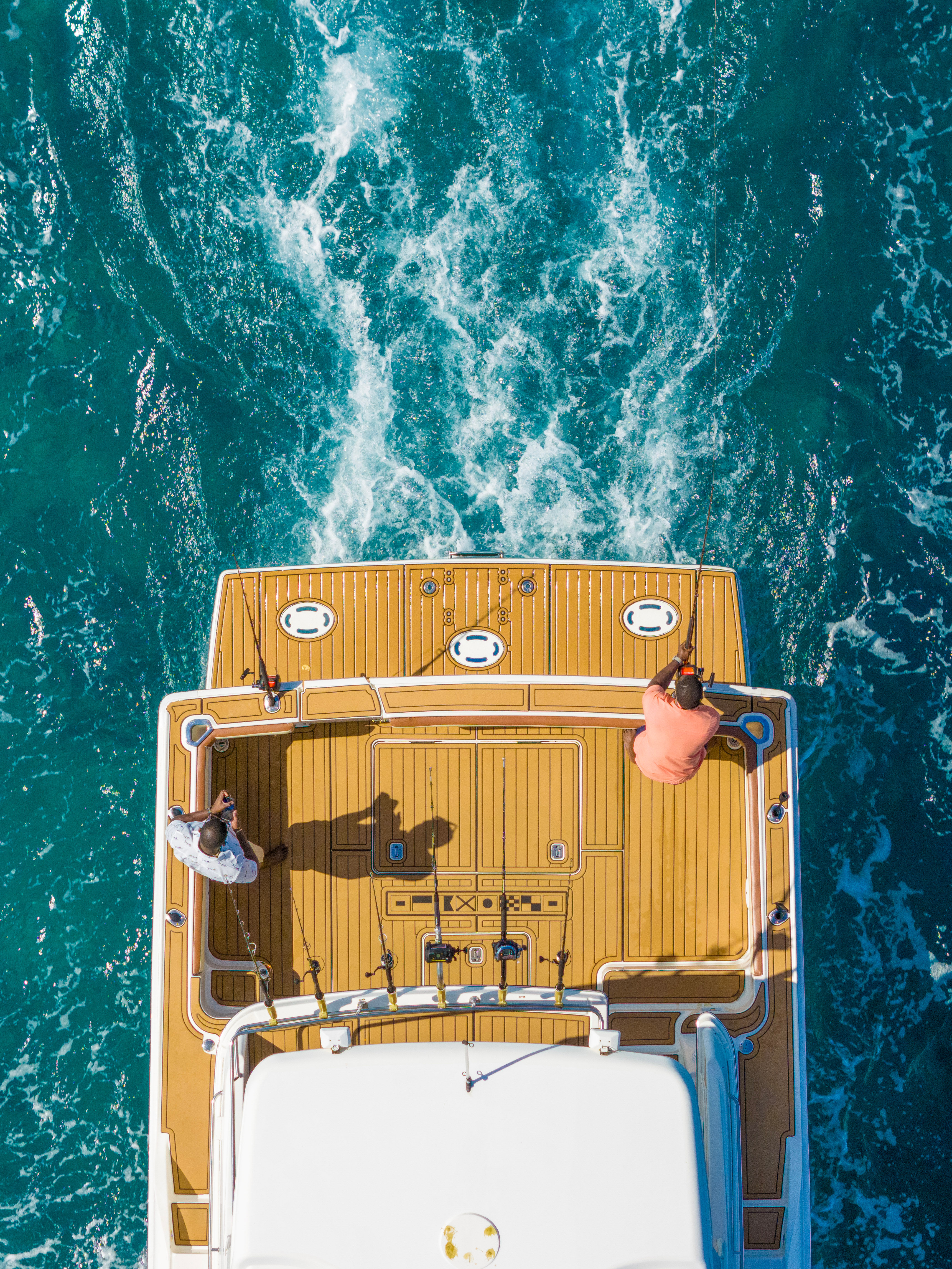 Aerial view of two anglers on a yacht’s teak deck fishing over turquoise ocean wake — offshore boating and sport-fishing scene.