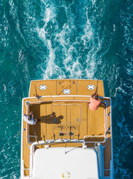 Aerial view of two anglers on a yacht’s teak deck fishing over turquoise ocean wake — offshore boating and sport-fishing scene.