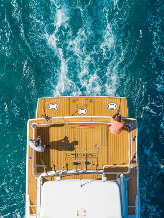 Aerial view of two anglers on a yacht’s teak deck fishing over turquoise ocean wake — offshore boating and sport-fishing scene.
