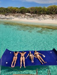 Four friends sunbathing on a large blue floating mat over crystal-clear turquoise water beside a white-sand tropical beach with trees