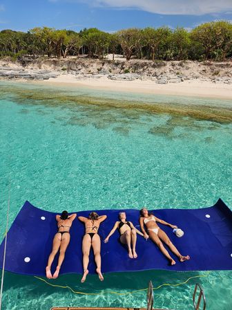 Four friends sunbathing on a large blue floating mat over crystal-clear turquoise water beside a white-sand tropical beach with trees