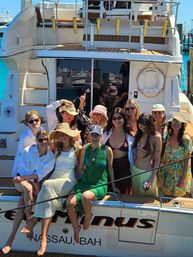 Group of friends laughing on the stern of a yacht in Nassau, Bahamas — sun hats, swimsuits and summer dresses with bright turquoise water and cruise ships reflected in the boat windows.