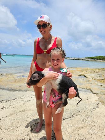 Smiling woman in a red bikini and a girl holding a black-and-white piglet on a sunny tropical beach with turquoise water and rocky shoreline.