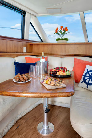Cozy yacht dining nook with wooden table set with wine in an ice bucket, empty glasses, skewers and fruit platter, nautical pillows and blue-sky windows