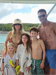 Family of five smiling on a boat in turquoise tropical waters — parents in swimwear and sunglasses, three kids in beach cover-ups and swim trunks holding snacks with a green island shoreline in the background.