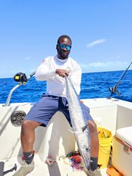 Offshore angler on a white boat holding a long silver wahoo (kingfish) with bright blue ocean and sky, sportfishing rods and gear visible.